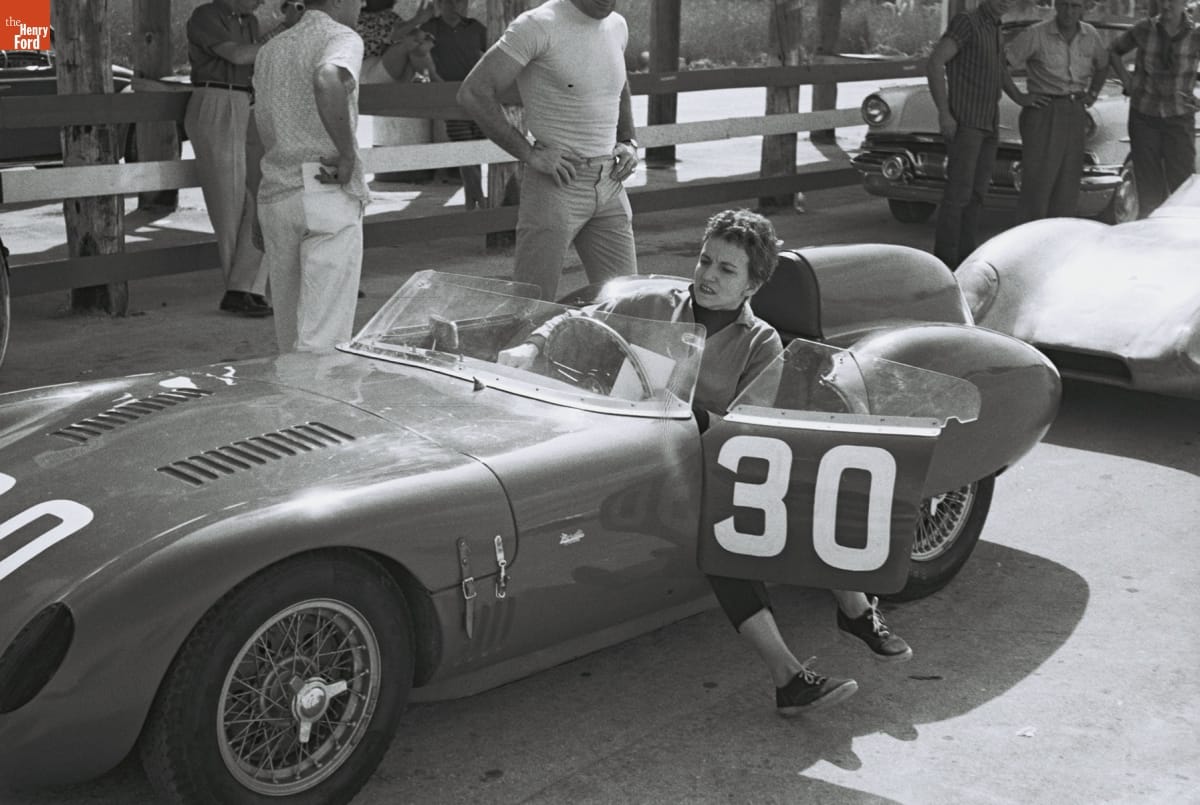 Woman sits in race car with feet dangling out open door; other people and cars in background