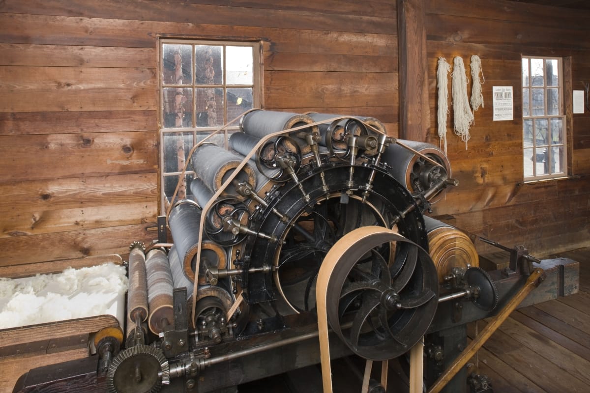 Machine with multiple rollers arranged in an arch shape with a conveyor belt filled with wool leading up to it