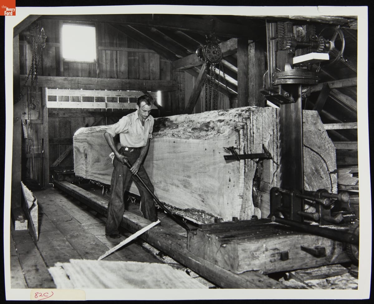 Black-and-white photo of man working next to very large tree trunk mounted on a machine in a wooden building