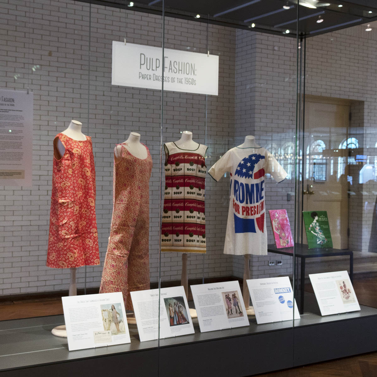 Glass case with four dress forms, each containing a garment, labels in front and additional artifacts on a low table