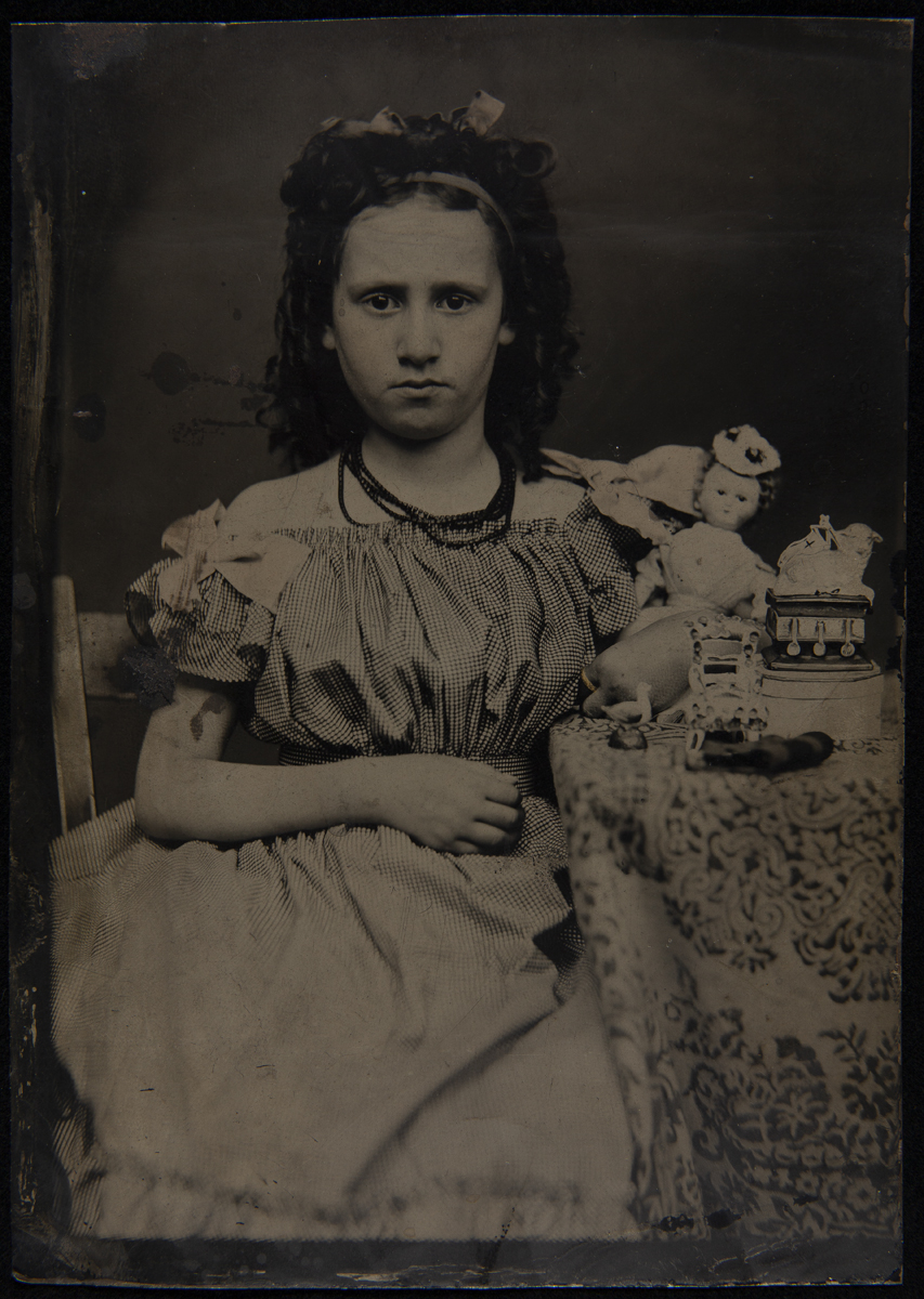Girl in necklace and dress sitting by a table covered with a tablecloth with toys on top
