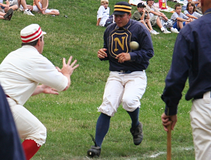Baseball player runs toward a plate as a ball is thrown toward the baseman and spectators look on