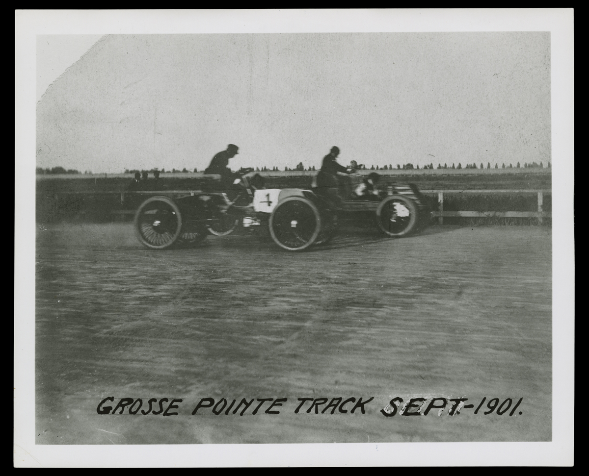 Two minimalistic early cars with a driver in each on dirt track with fence and grass in background