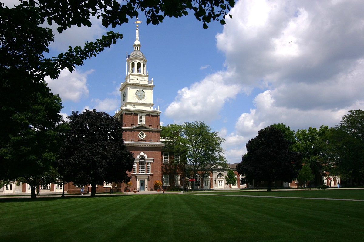 Brick building with tall clocktower, seen across a large grass lawn with a few trees