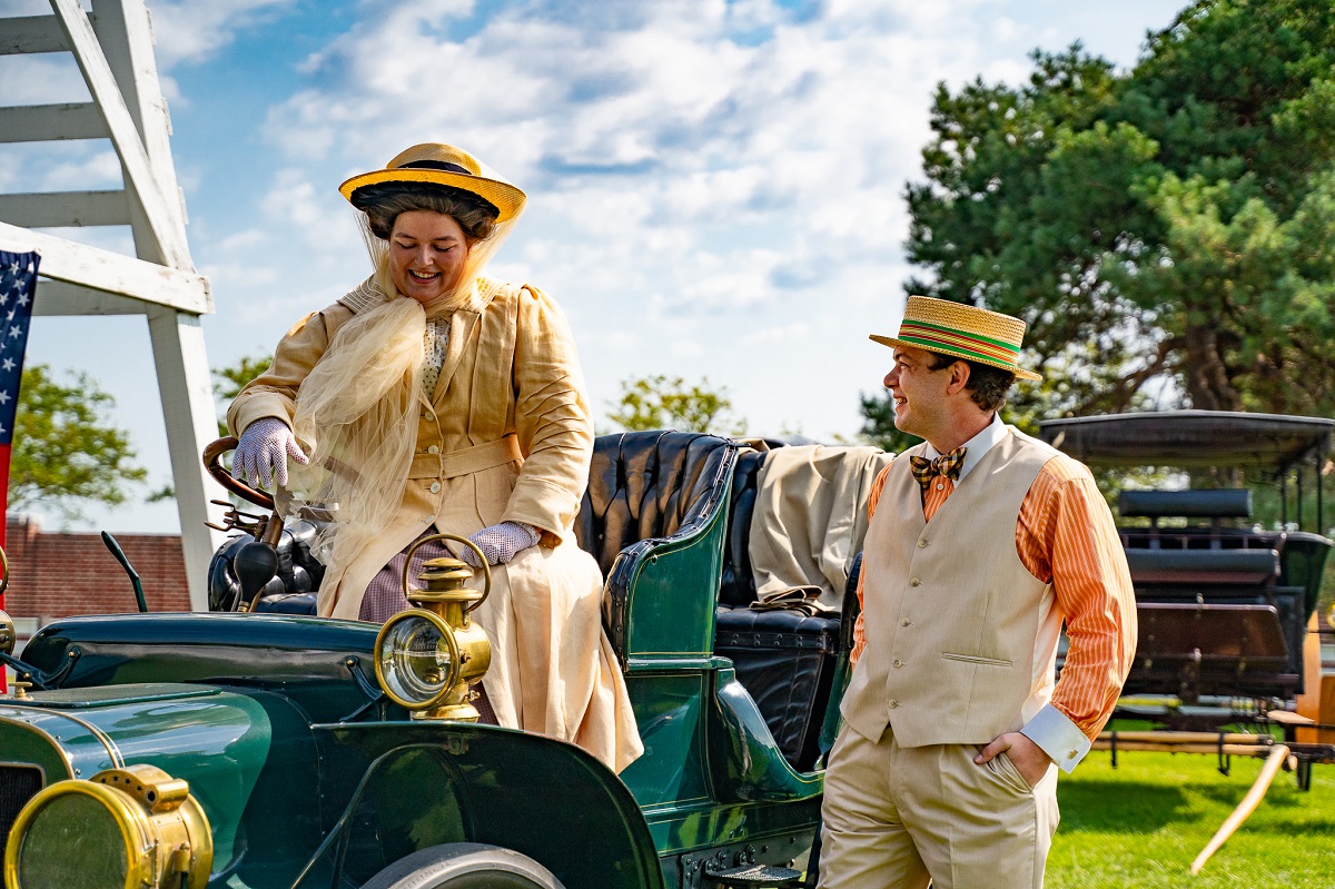 Woman in yellow coat and hat sits behind the wheel of an open car as a man in a vest and straw boater hat stands nearby