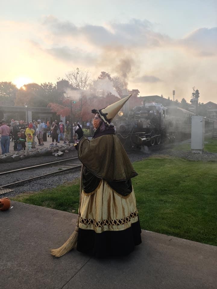 A witch and the Hallowe’en Express welcome guests to Hallowe’en in Greenfield Village in 2020 Woman in witch costume in front of a locomotive and crowd of people