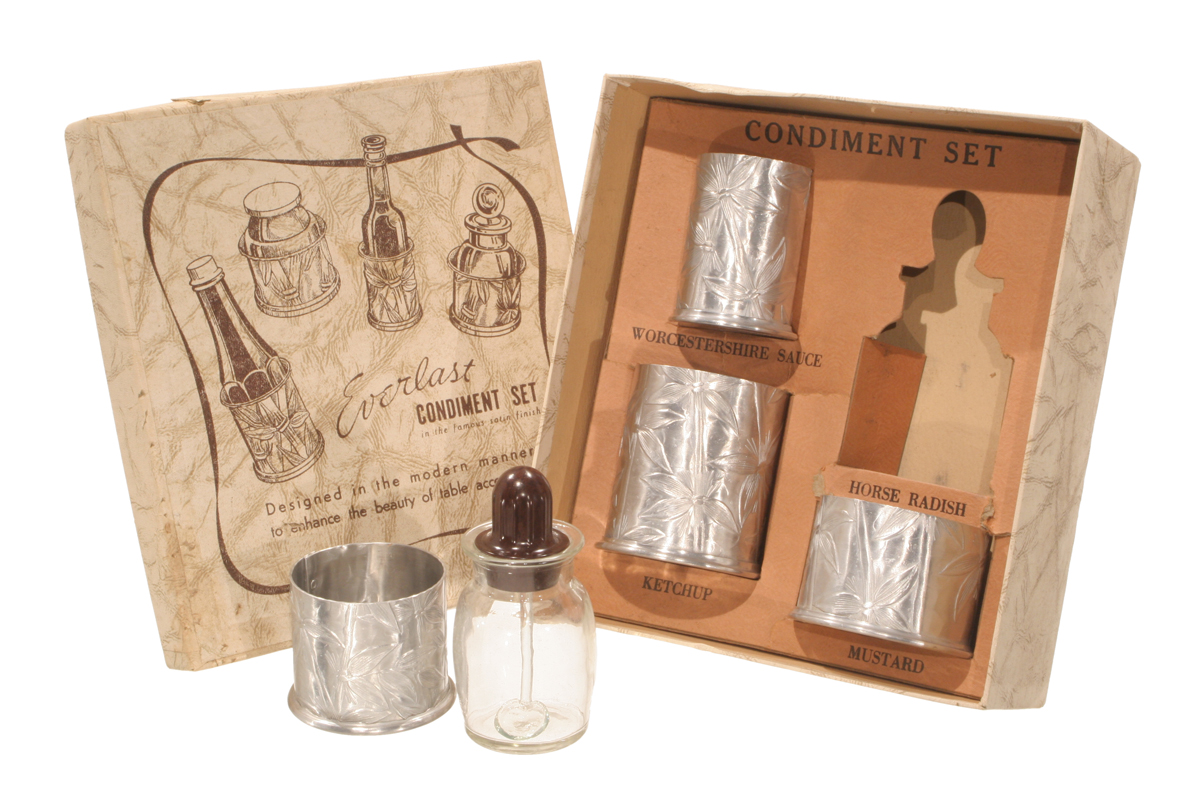 Box containing silver canisters; additional silver canister and glass in front; box lid with text and drawings of canisters nearby