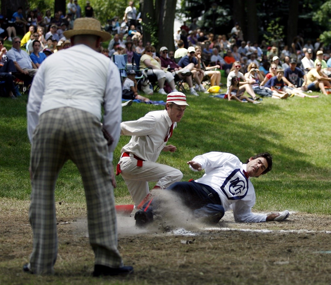 A historic base ball player slides onto a base while other players and a modern crowd looks on