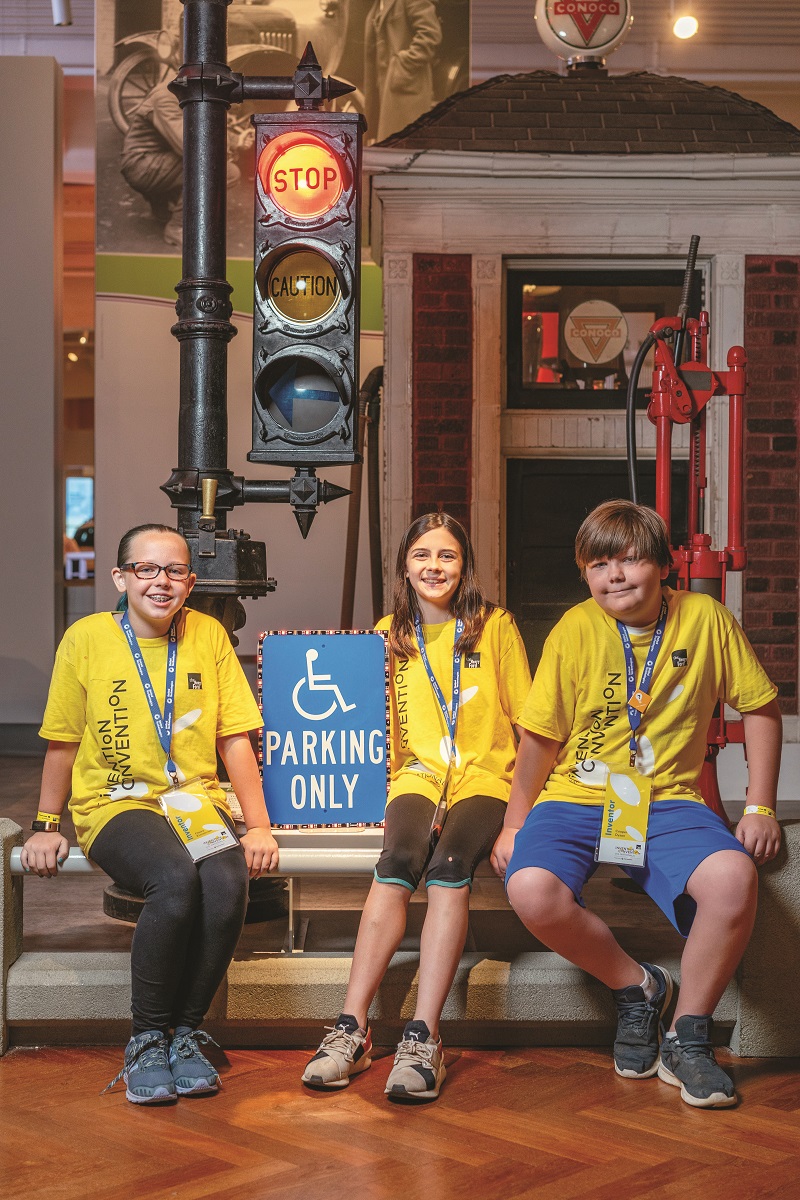Three children in yellow t-shirts sit in front of a old-fashioned traffic light and small brick gatehouse in a larger building; one holds a blue and white "handicapped" parking sign