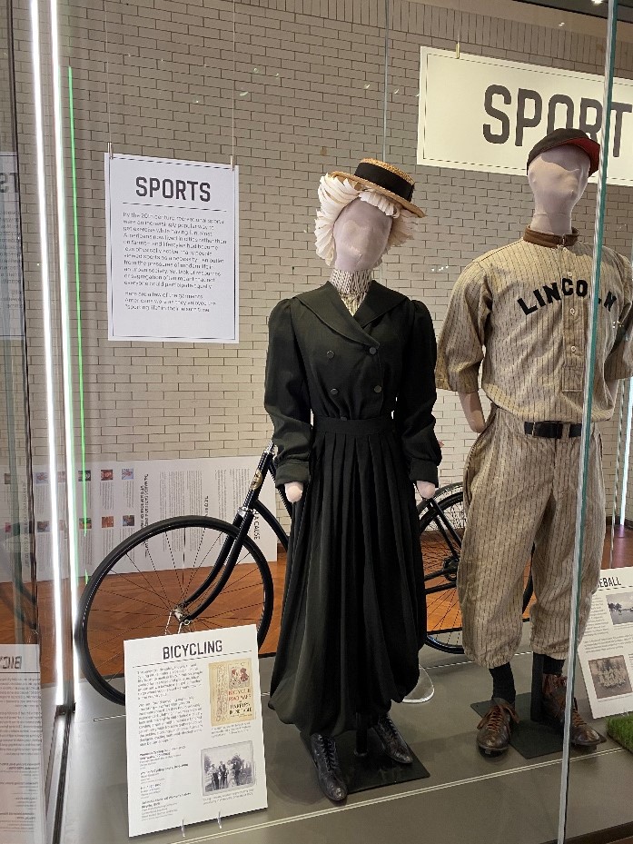 Mannequin wearing dress and paper wig topped with straw boater hat in display case with other items and text panels