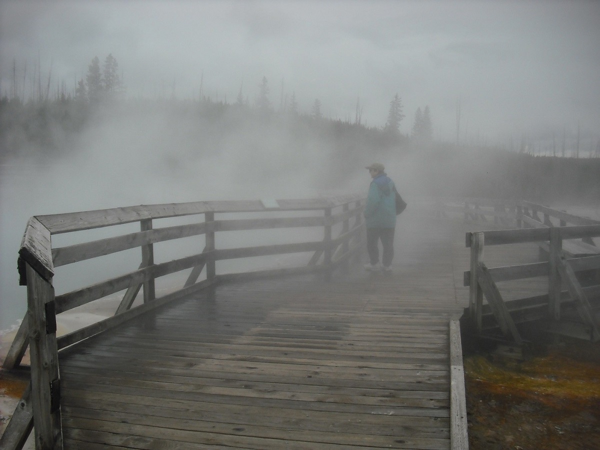 Woman stands on wooden boardwalk among steam, mist, or fog with trees visible in the background