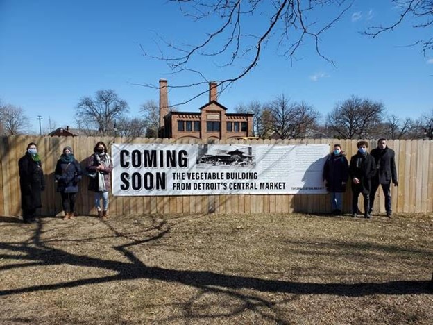 Debra Reid, Kathleen Brown, Ayana Curran-Howes, Laurel Fricker, Antonello Mastronardi, and Jim Johnson, standing outside the fenced off area where the Vegetable Building is being erected Six people pose in front of a wooden fence on either side of a sign reading "Coming Soon: The Vegetable Building from Detroit's Central Market"