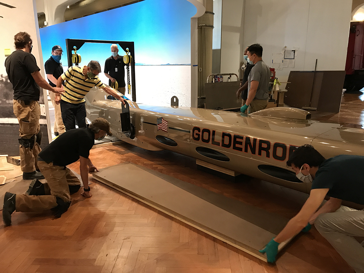 Group of men wearing facemasks stand and kneel around a long, low, torpedo-shaped golden car
