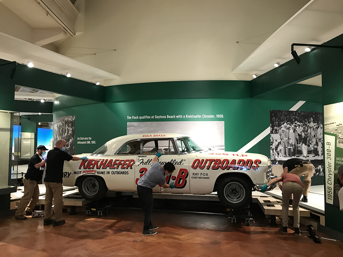 White car with bold red text along the side with several people guiding it into place in an exhibit
