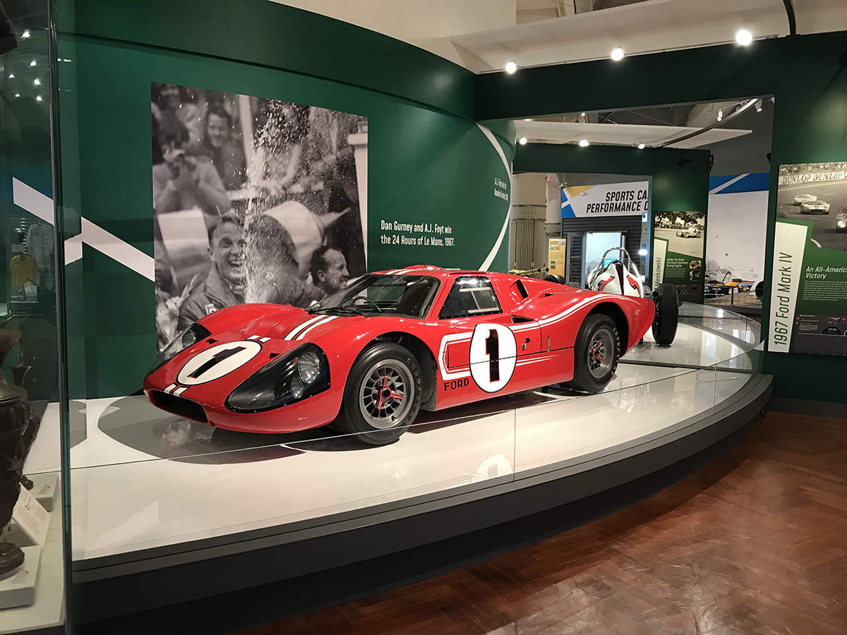 Low red race car with number "1" in a circle on side sits on a platform in front of a green backdrop with a large photo and text