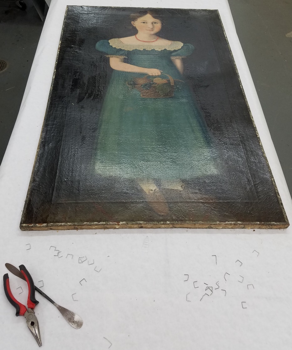 Painting of girl in blue dress, laid flat on table with pliers, metal spatula, and staples on table at the end