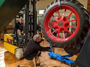 Large tire dangling from forklift, being guided by a man kneeling on the ground