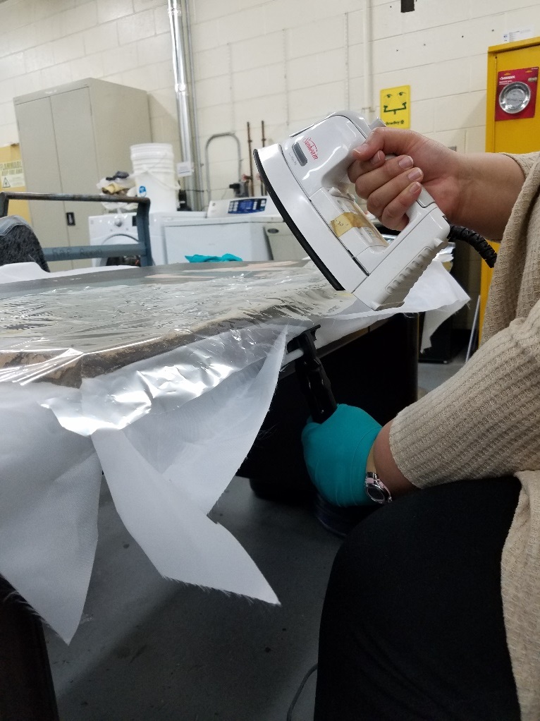 Person wearing gloves holds an iron to a flat item on a table in a lab