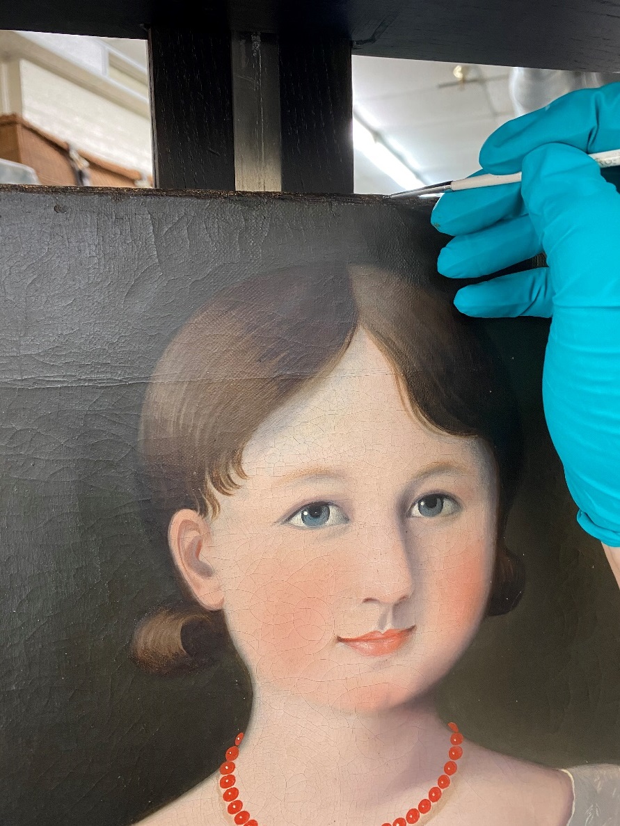 Blue-gloved hand holds a paintbrush at the top of a painting of a girl's head