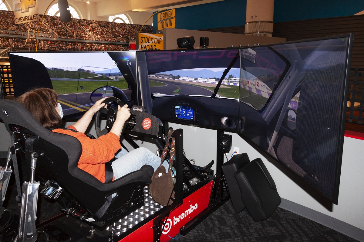A person sits in a car simulator in front of three large screens displaying a race track