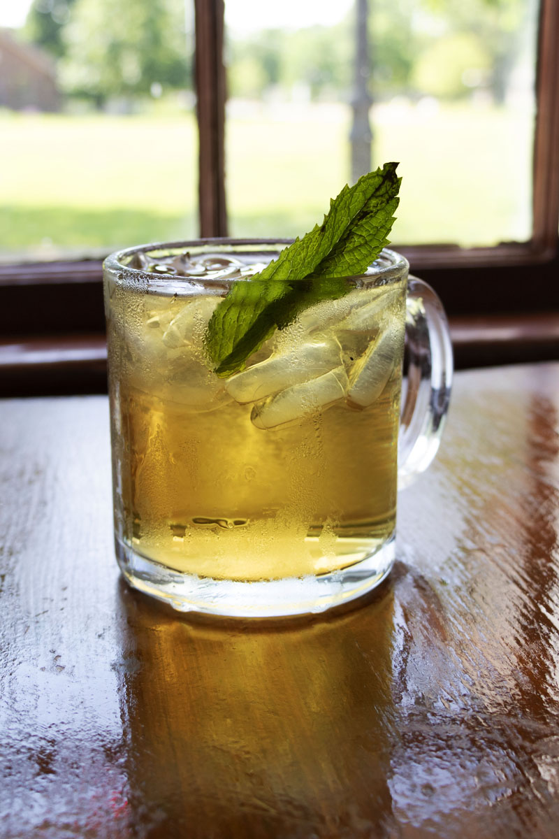 Clear glass mug filled with ice and amber-colored liquid, garnished with a mint leaf, sitting on a wooden table in front of a window