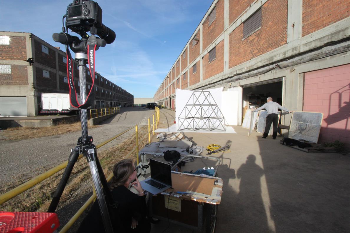 Camera on tripod in foreground pointing toward white paper with metal truss in front of it alongside a large red brick building