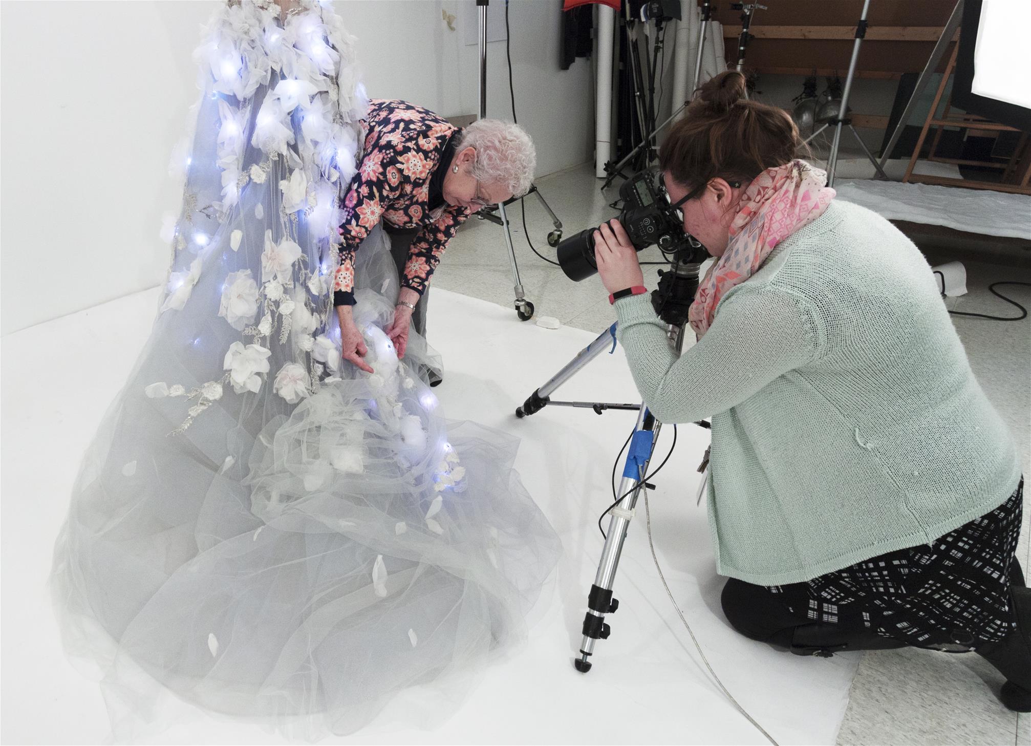 Woman in foreground looks through camera as a second woman holds up part of a dress skirt