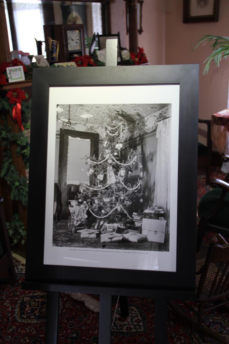 The original historic photo of the Wright family Christmas tree, displayed in the Wright Home during Holiday Nights in Greenfield Village. Large framed black-and-white photograph showing a Christmas tree on an easel in a room