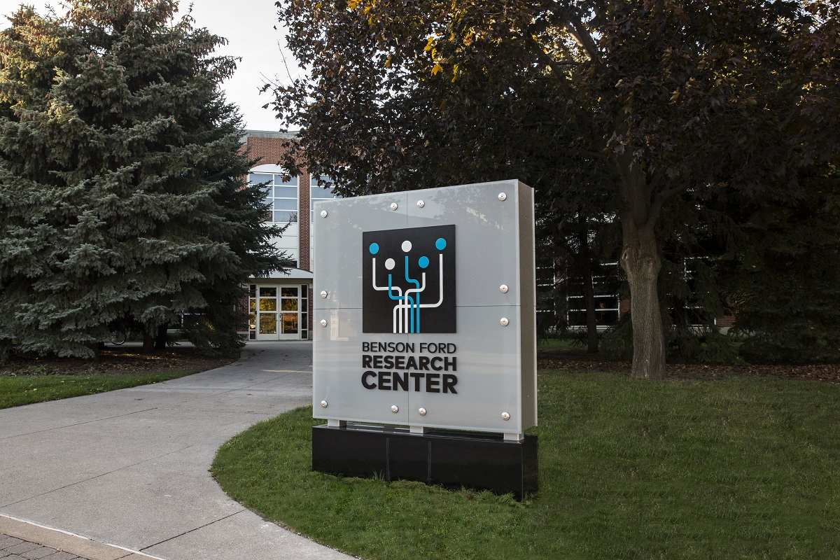 Photo of gray sign with text and logo on lawn next to curving sidewalk leading to a brick building