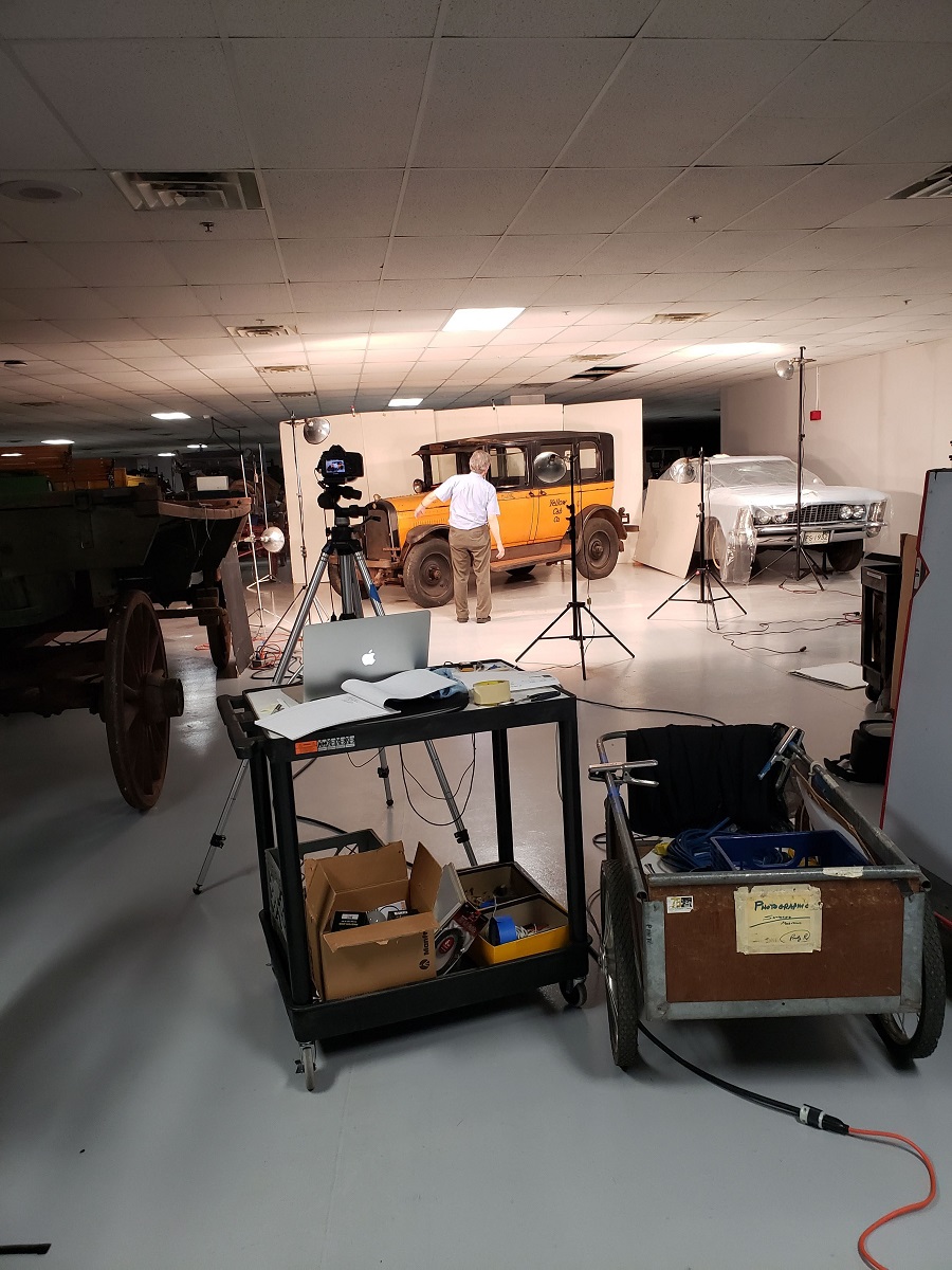 A yellow car sits in a cramped space among other vehicles in front of a white background, with carts of photo equipment, a camera on a tripod, and lights on stands in the foreground