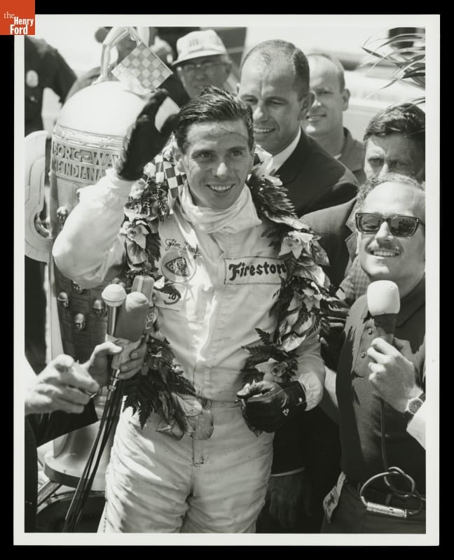 Man in jumpsuit and gloves with wreath around neck waves, surrounded by a crowd