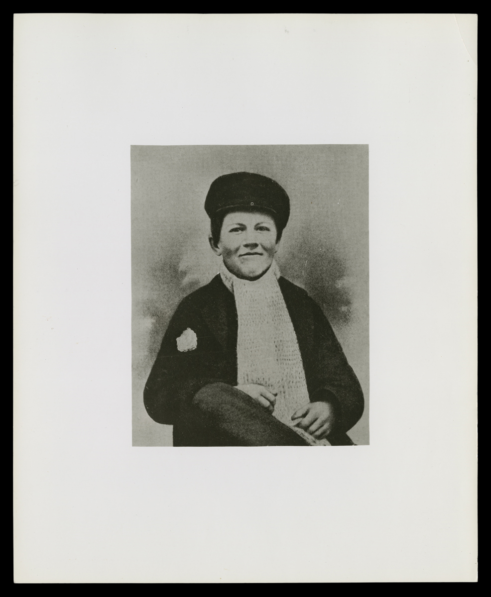 Black-and-white photo of seated young boy in hat, scarf, and jacket