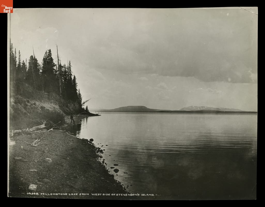 Black-and-white photo of a lake with a small shoreline with pine trees and a man fishing, with hills or mountains in the background