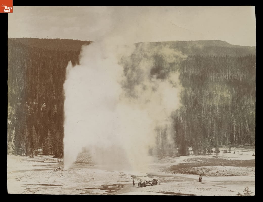 Black-and-white photo of geyser spewing water and steam into the air as people look on; vast pine forest in the background