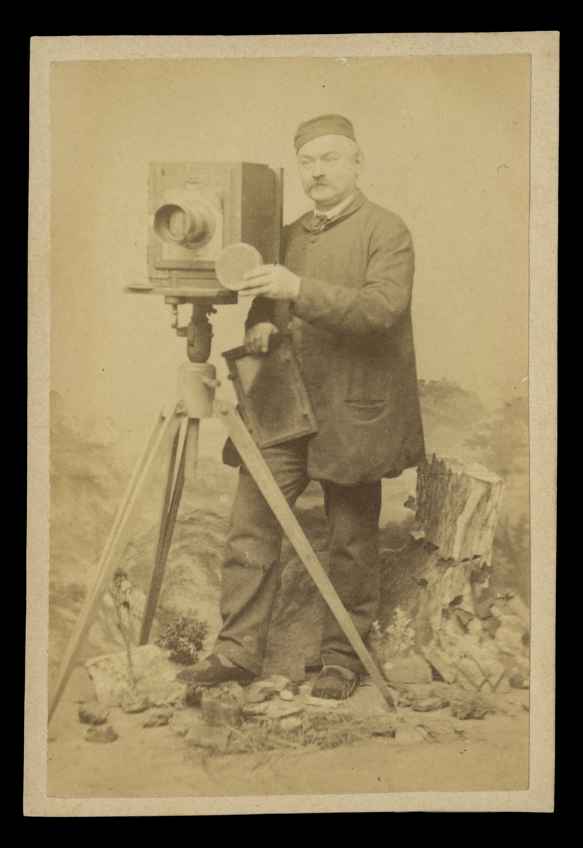 Studio Portrait of a Photographer with His Camera, circa 1870 Man in smock and cap standing behind camera on tripod