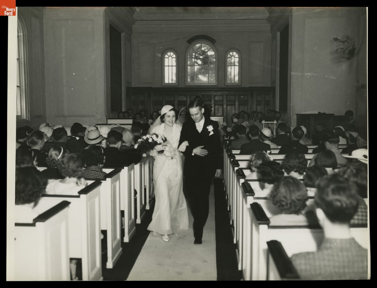First Wedding Held in Martha-Mary Chapel in Greenfield Village, 1935 Bride and groom, arm in arm, walking down a church aisle past pews full of people sitting facing away from the camera