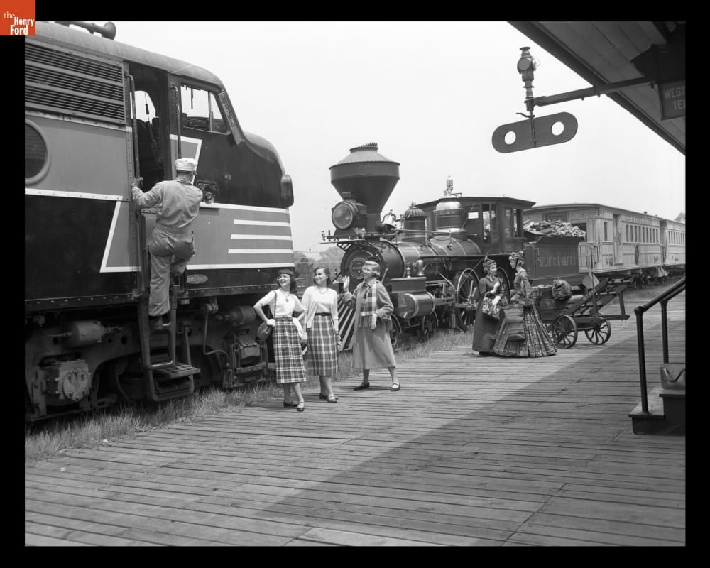 New York Central Diesel Locomotive Facing "Sam Hill" Steam Locomotive in Greenfield Village, May 1953 Two locomotives face each other on a track, with women in period clothing on the platform in front of them