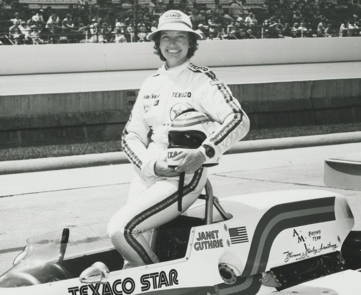 Woman in racing jumpsuit holding helmet stands in race car on track with stands of people behind her