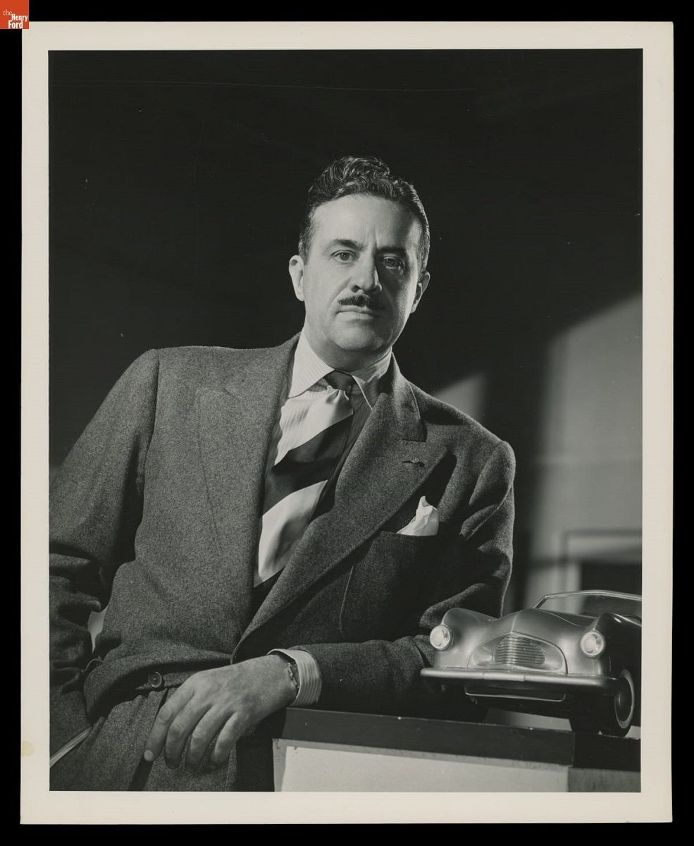 Man with mustache wearing suit and striped tie leans on a surface holding a small car model