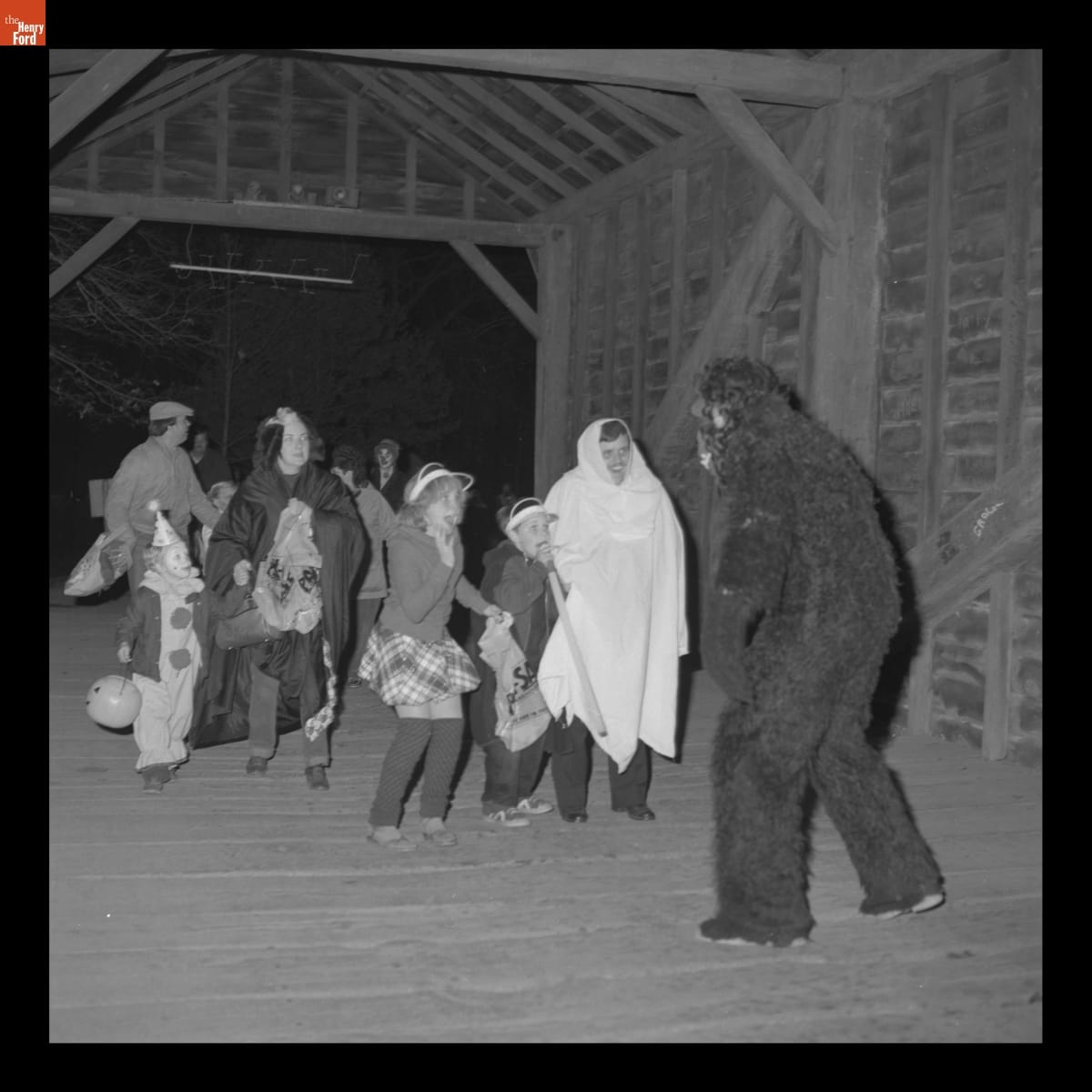"Gorilla" on Ackley Covered Bridge during Halloween in Greenfield Village, October 1982 Person in a gorilla costume in front of adults and children, many in costume, in a wooden covered bridge