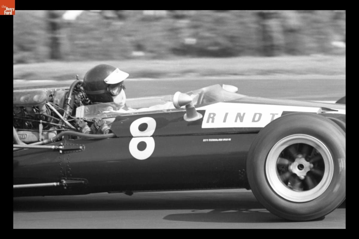 Partial side view of racecar with text "8" and "RINDT" on side; person inside in helmet, goggles, and mouth covering