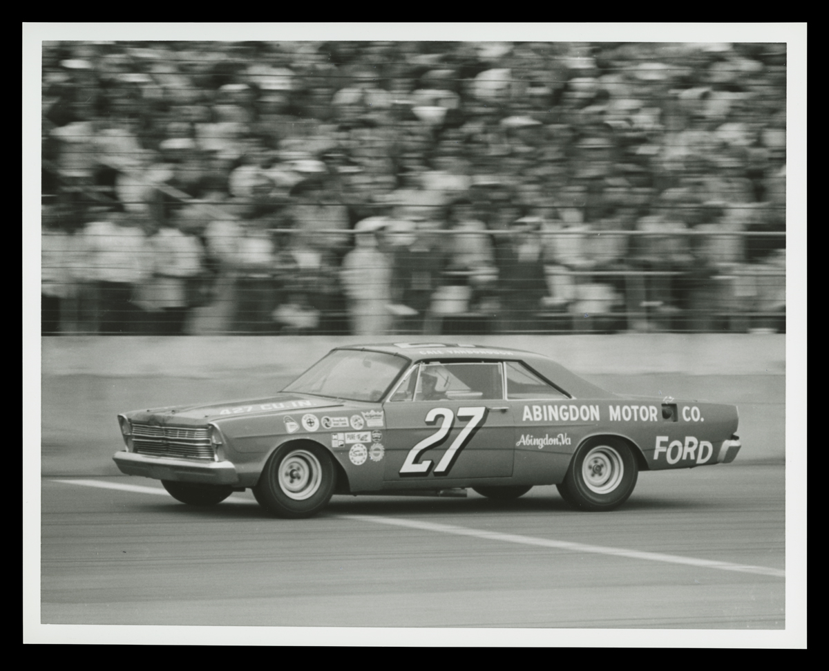 Car, with large number 27 on door, in front of blurred stands full of people