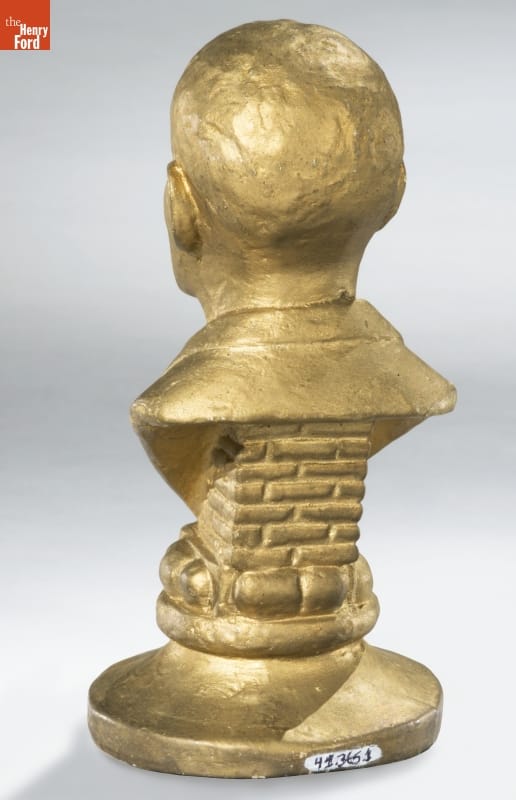 Back of gold sculpture of man's head and partial torso on a base designed to look like bricks