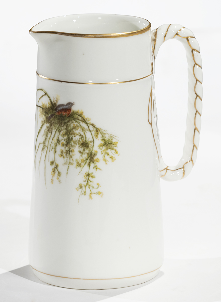 White pitcher with gold piping on handle and at top and painted illustration of bird atop vegetation 