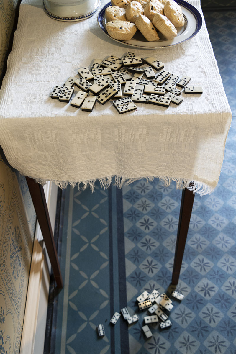 End of table covered with cloth with dominos and plate of scones on it; additional dominos on patterned floor below 
