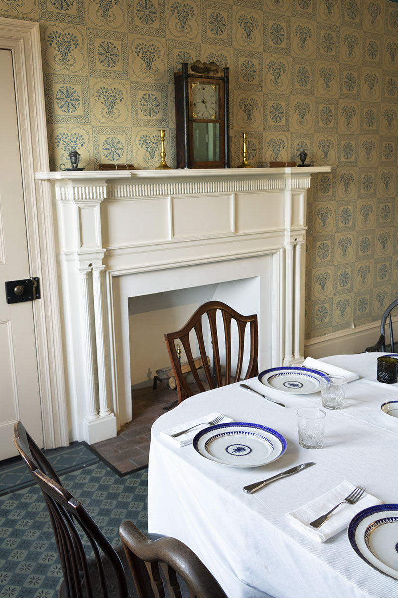 Corner of set table with chairs; fireplace with mantel behind and patterned wallpaper on walls