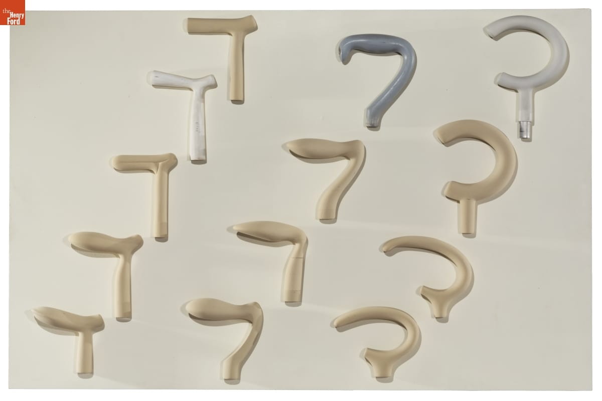 Board with different styles and shapes of cane handles mounted on it--mostly gold but also silver and gray colored