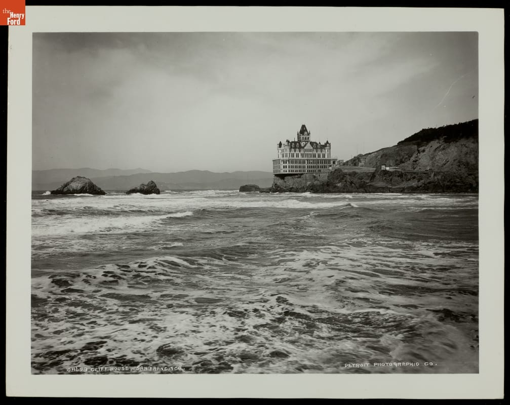 Black-and-white photo of large building at the edge of bluffs over a body of water 