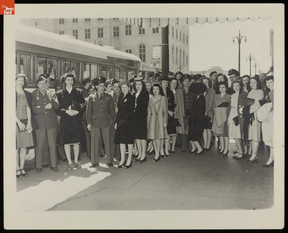 Group of men in military uniforms and women in dresses stand in front of a bus, with buildings and streetlights in the background
