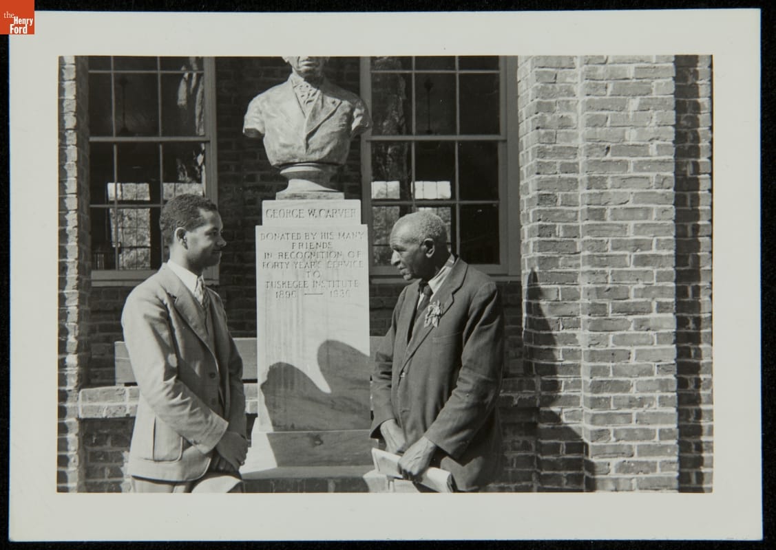 Black-and-white photograph of two men in suits standing on either side of a sculpture of a man's head and torso above a tall base, all in front of a brick building with windows 